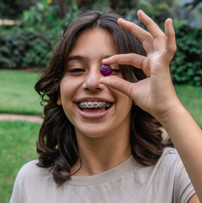 A smiling girl with braces holding a purple gummy in front of her eye.