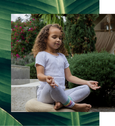 A smiling child with her eyes closed, meditating outdoors. The image is framed by large green leaves.
