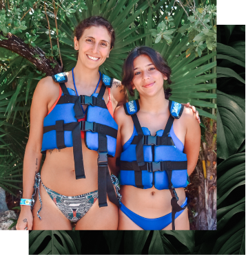 A smiling woman and girl in life vests, standing together in front of green leaves.